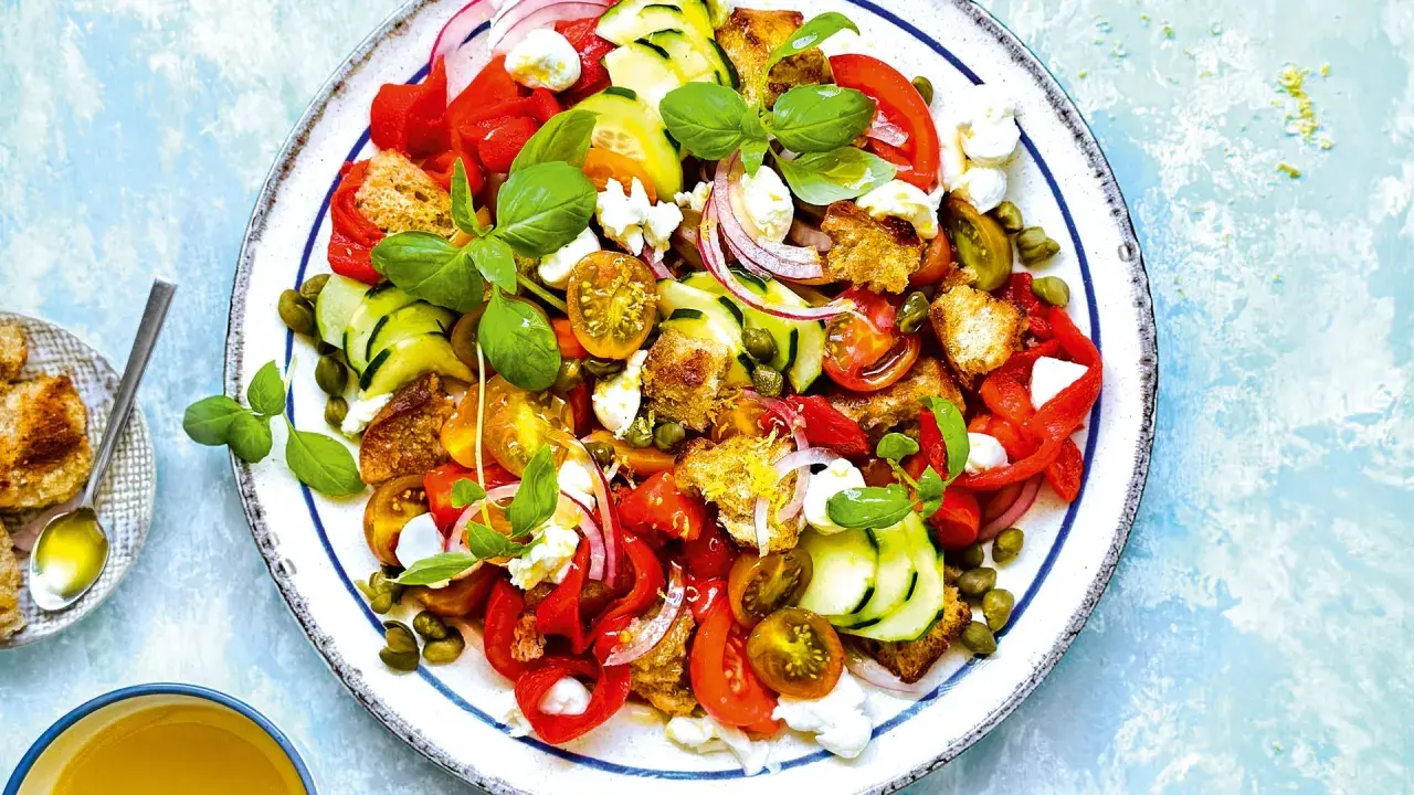 Ensalada de burrata vibrante con tomates, pepino, pimientos rojos, pan tostado y alcaparras, adornada con albahaca fresca.