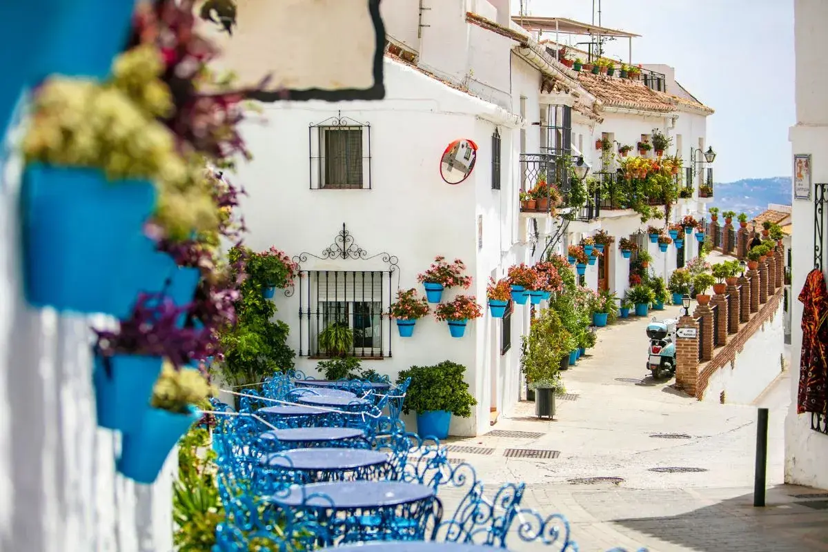 Callejuela encantadora en uno de los pueblos costeros de Málaga, con fachadas blancas, macetas azules y mesas de terraza.