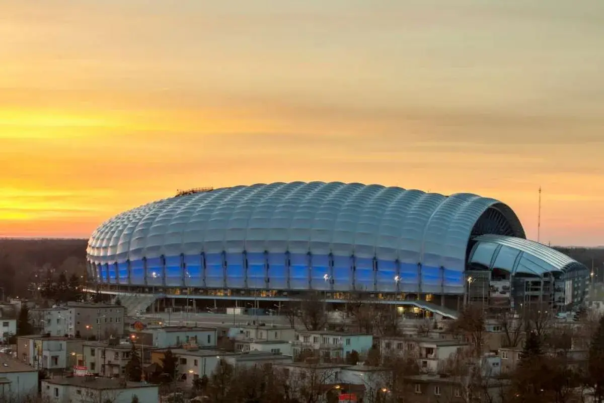 Stadion miejski w Poznaniu o zachodzie słońca, z niebieskimi światłami i charakterystycznym, falującym dachem.
