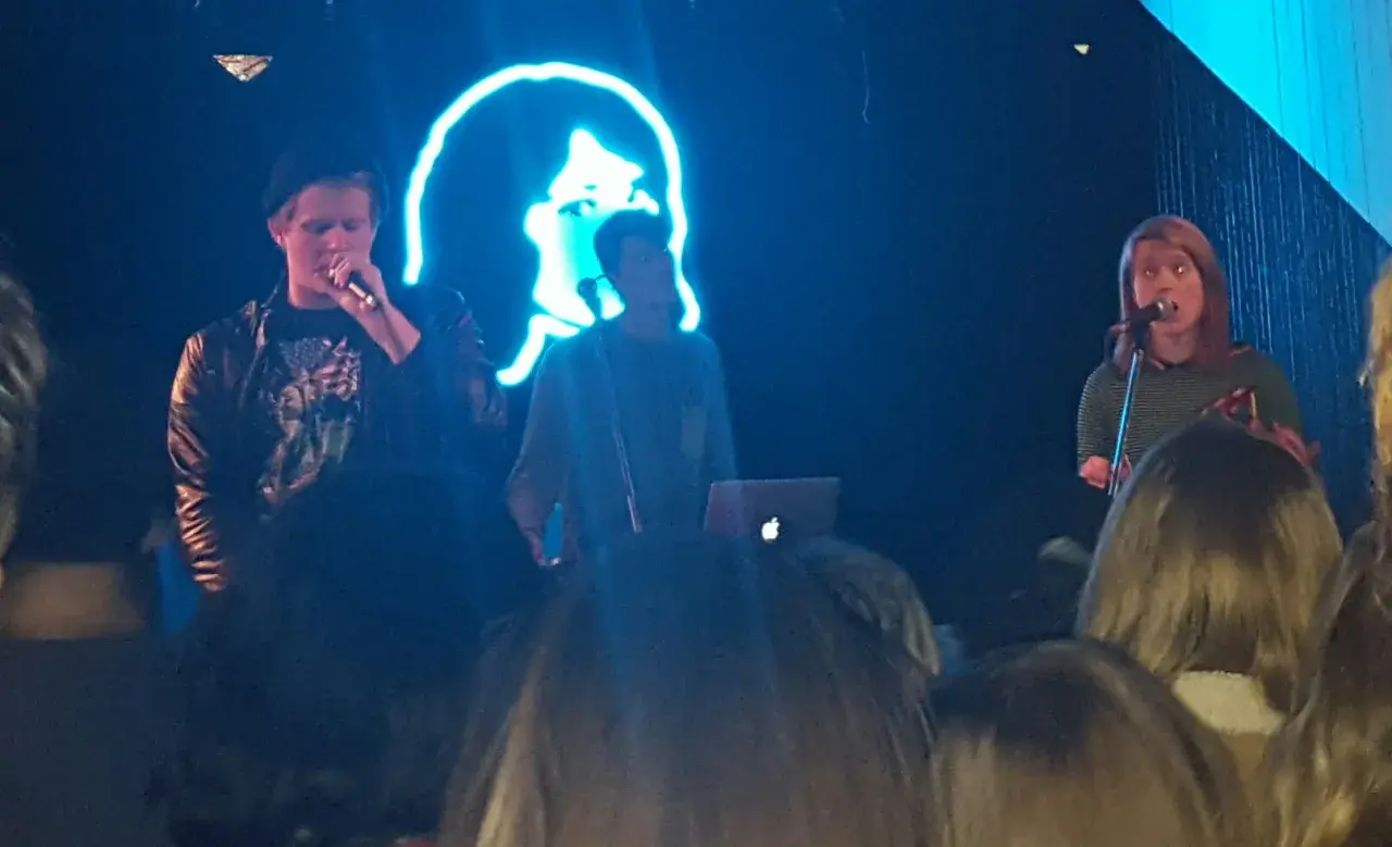 Koncert zespołu tv girl. Na scenie trójka artystów, w tle neonowa podobizna twarzy.