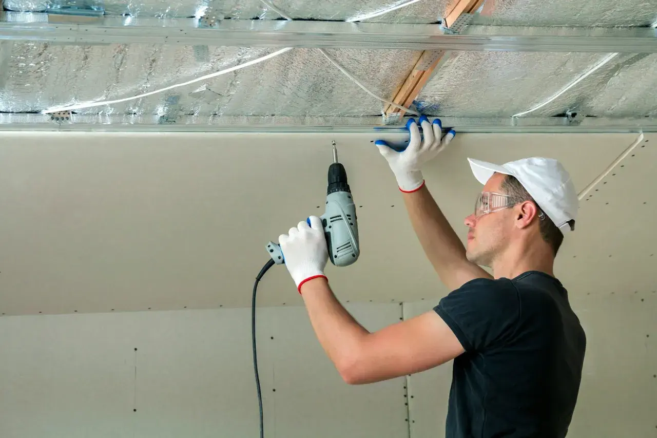 Un ouvrier installe des isolants minces sous un faux plafond en plaques de plâtre, utilisant une perceuse pour fixer les éléments.