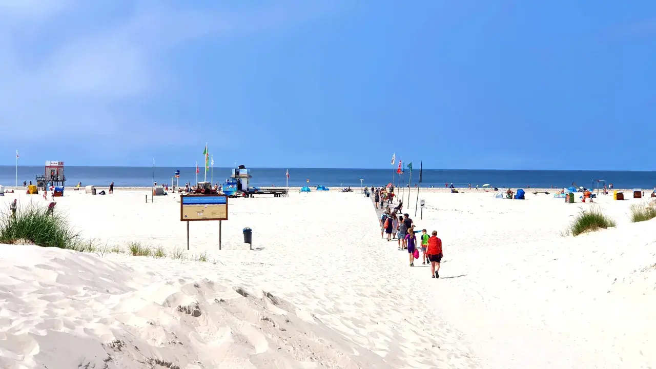 Menschen spazieren auf einem Holzsteg über den weiten, weißen nordsee strand. Der Himmel ist blau, das Meer ruhig.