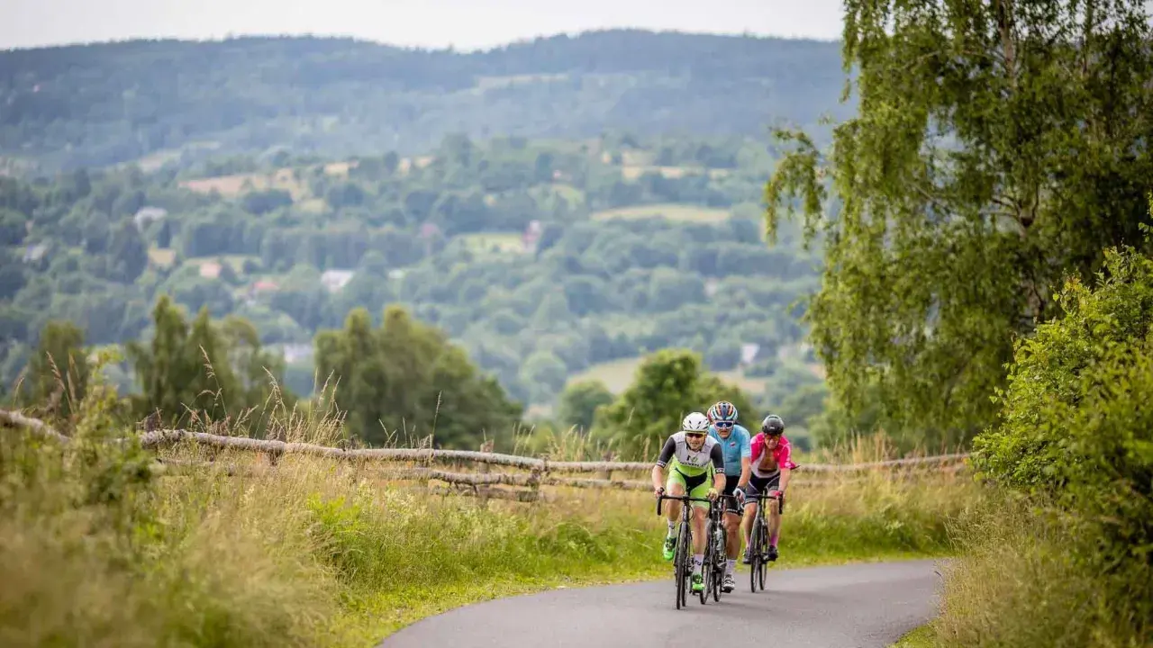 Trzech rowerzystów jedzie po asfaltowej drodze wśród zielonych wzgórz.