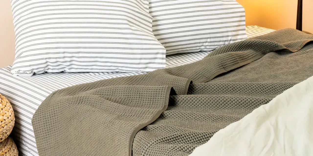 A textured, grey blanket is draped at the end of a bed, over striped pillows and sheets.