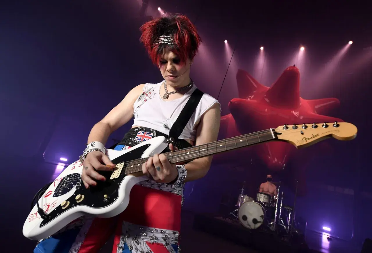 Yungblud gra na gitarze podczas koncertu w Warszawie. W tle czerwona, dmuchana instalacja i perkusista.