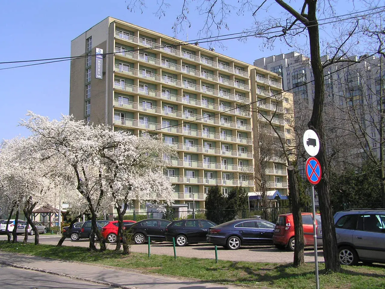 Hotel Felix Warszawa w otoczeniu kwitnących drzew. Widać parking z samochodami i budynek hotelu z balkonami.