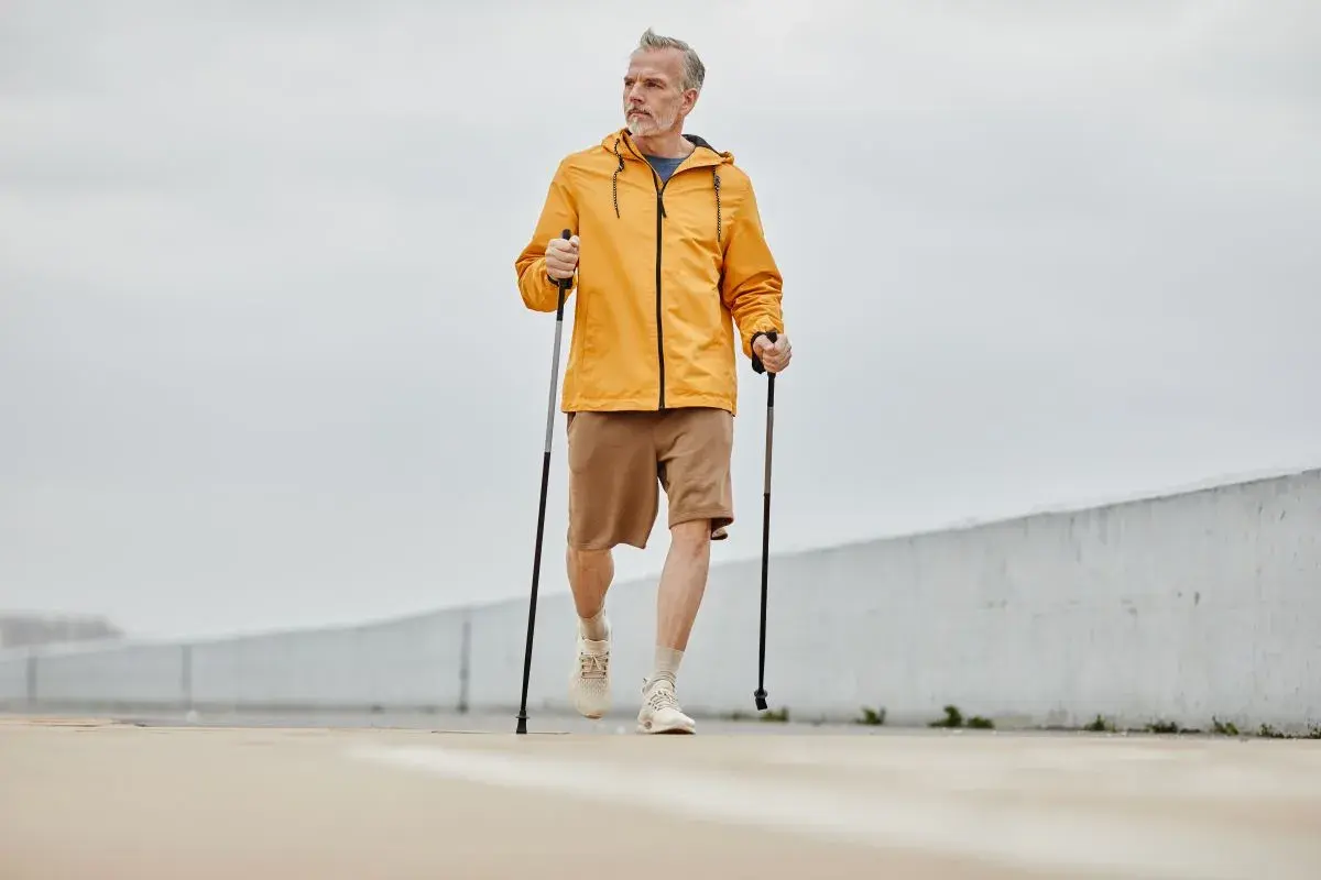Mężczyzna w żółtej kurtce i brązowych spodenkach wykonuje rozgrzewkę przed nordic walking, trzymając kije.