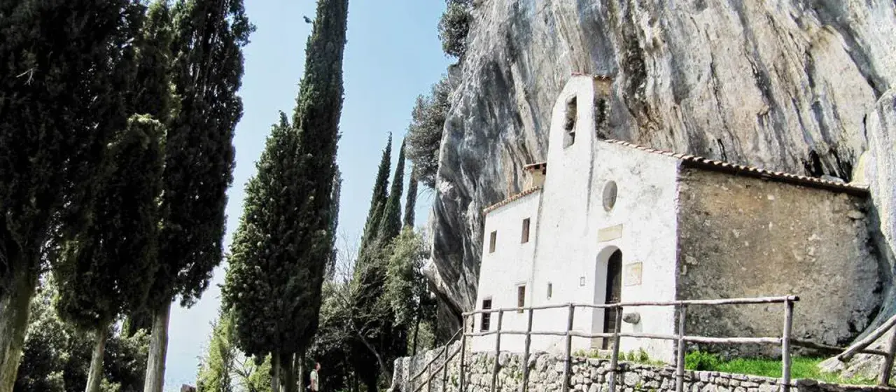 Piccolo eremo di San Valentino incastonato nella roccia, circondato da alti cipressi e un cielo azzurro.