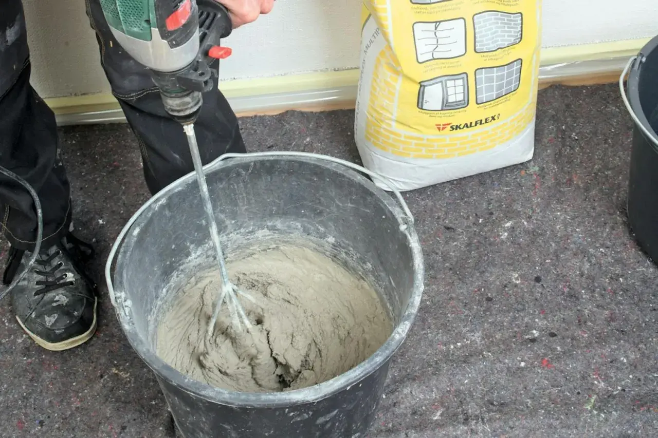 Personen använder en borrmaskin för att blanda betong i en hink. En gul säck med Skalflex-märkning står bredvid.
