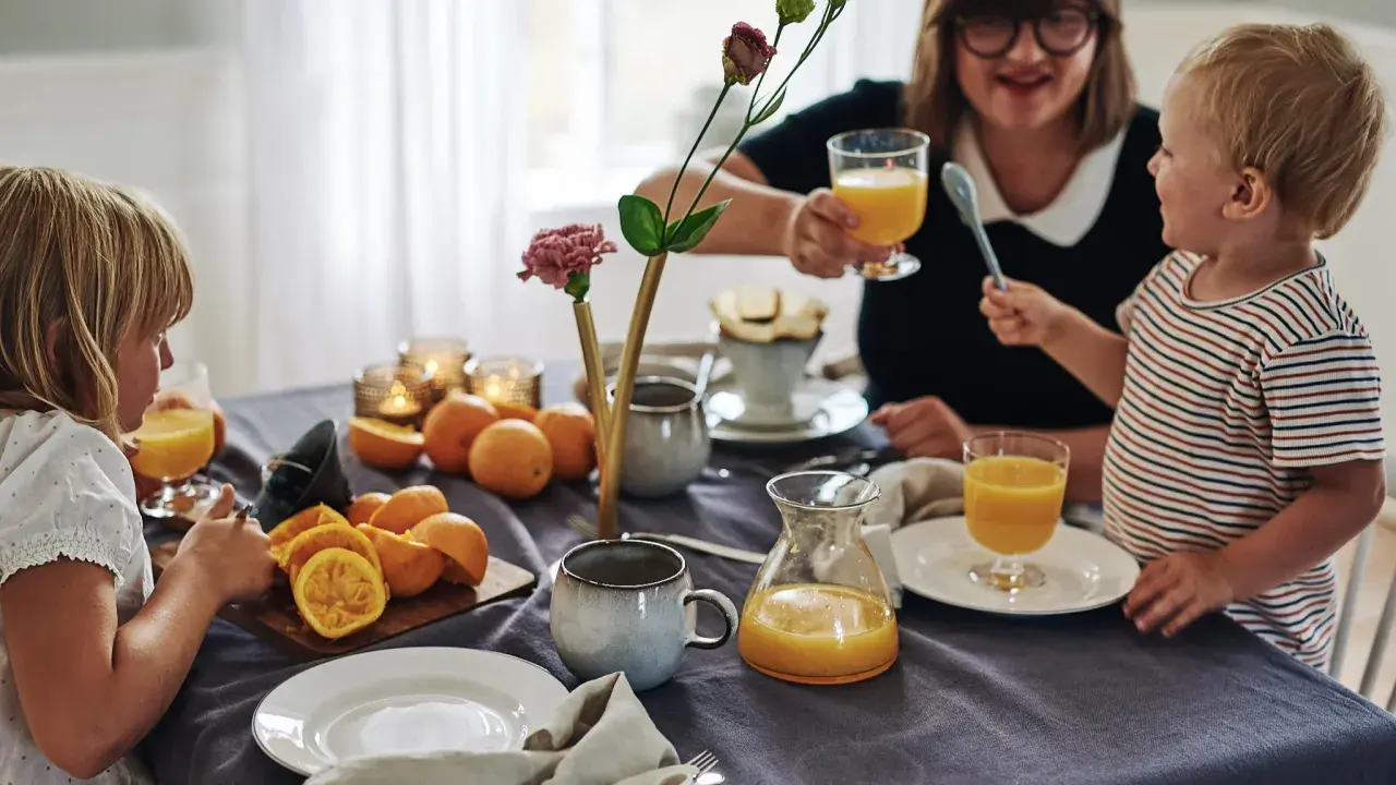 Familjen samlas kring frukostbordet med apelsinjuice och frukt. Duken är av linne, redo att stryka för nästa måltid.