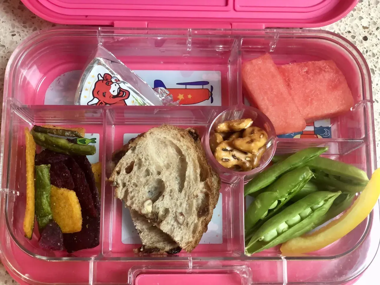 A pink bento box with watermelon, peas, pretzels, bread, and veggie sticks. Add an ice pack to keep this bento box cold.