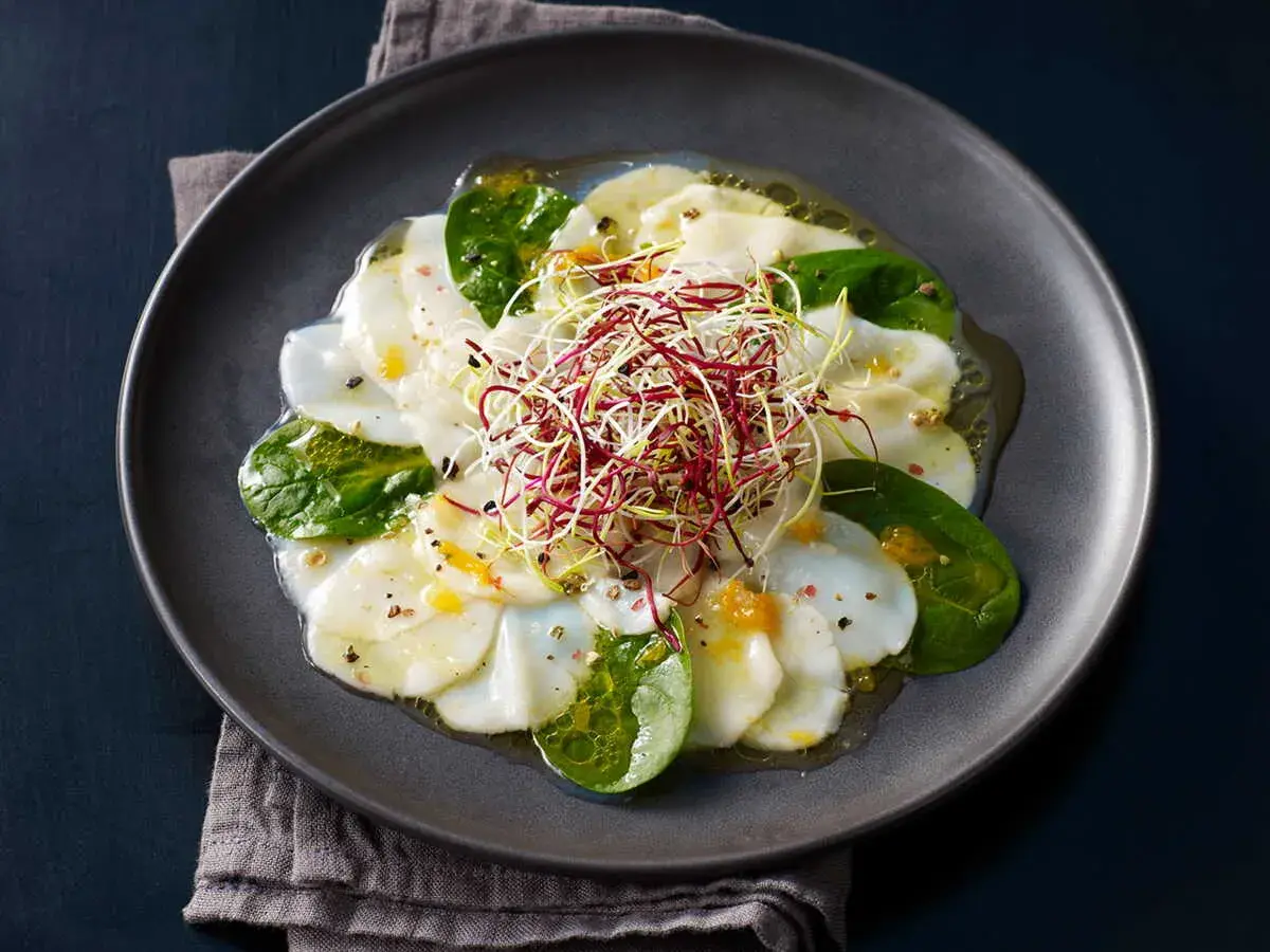 Carpaccio de Saint-Jacques aux agrumes, garni de pousses croquantes et de feuilles d'épinard frais, arrosé d'une vinaigrette légère.