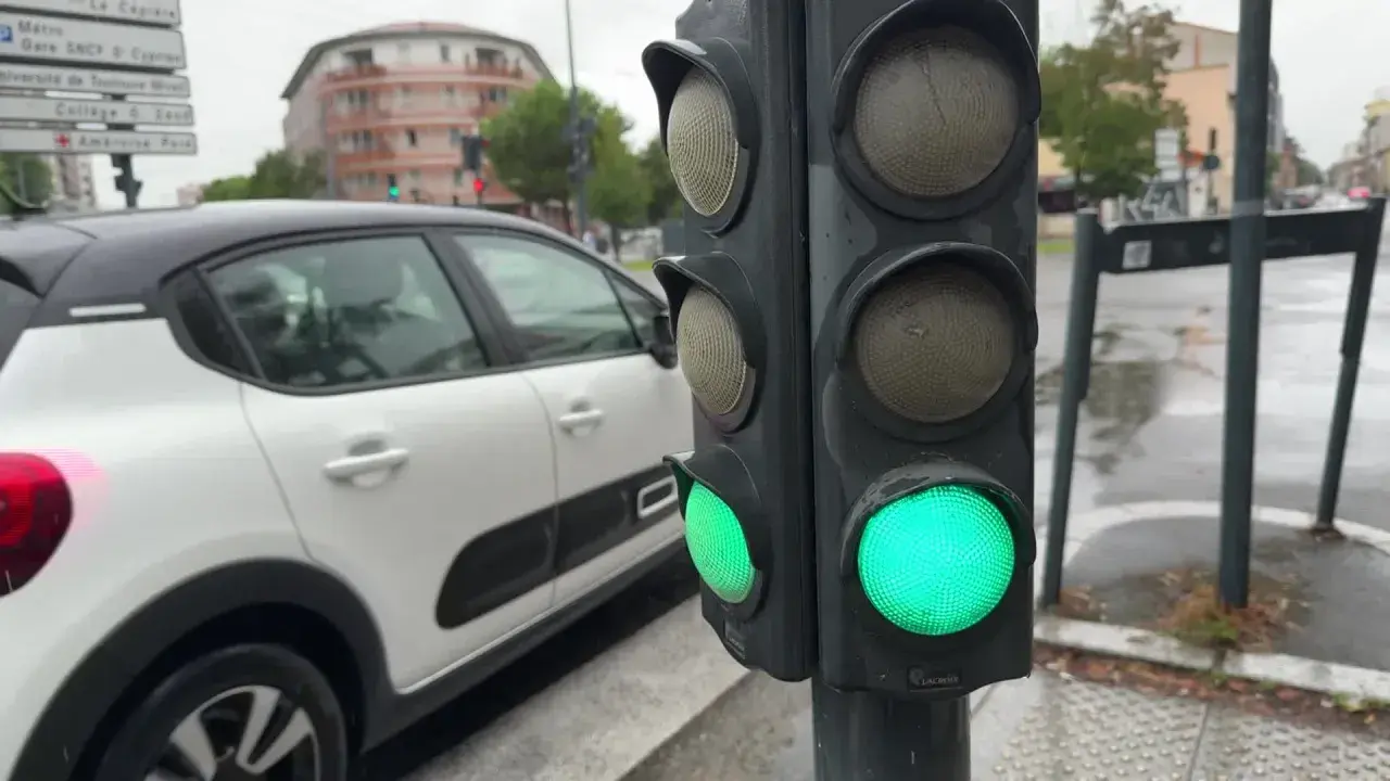 Un feu de circulation vert est allumé, indiquant que la circulation peut passer. Le gyrophare vert signification est la permission de continuer. Une voiture blanche est garée à proximité.