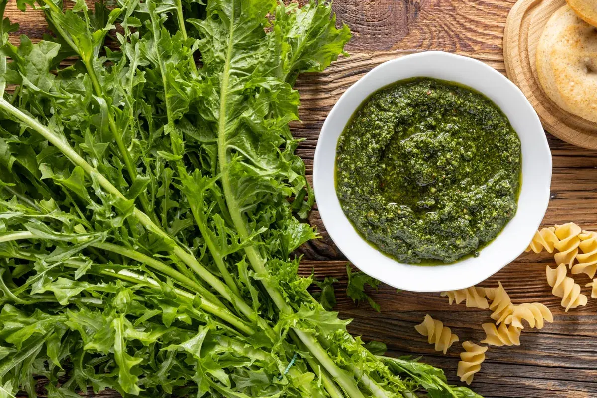 Un mazzo di **tarassaco foglie** fresche accanto a una ciotola di pesto verde brillante e pasta fusilli su un tavolo di legno rustico.