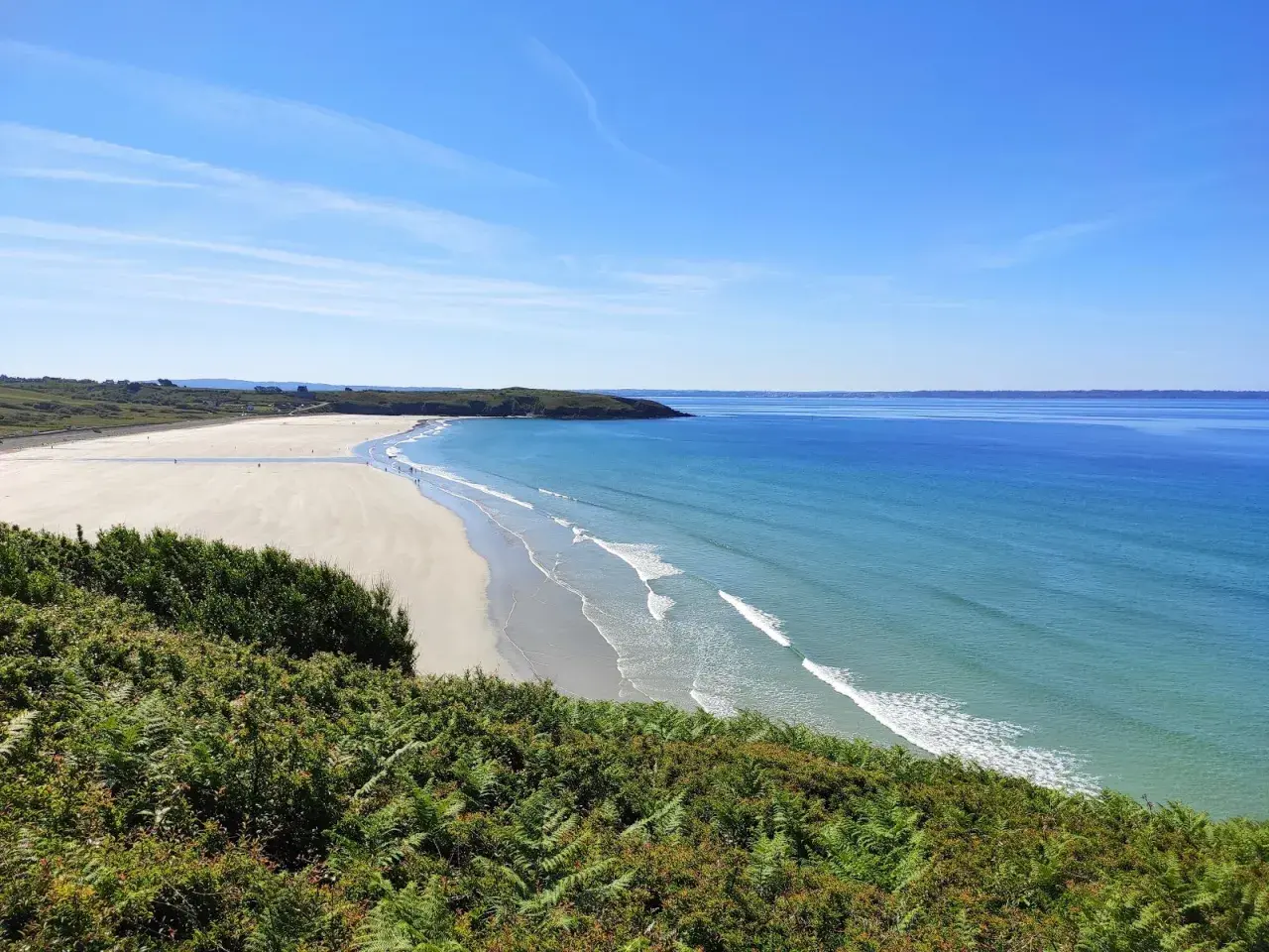 Vue panoramique d'une plage trez bellec, avec sable clair, mer bleue et végétation verdoyante au premier plan.