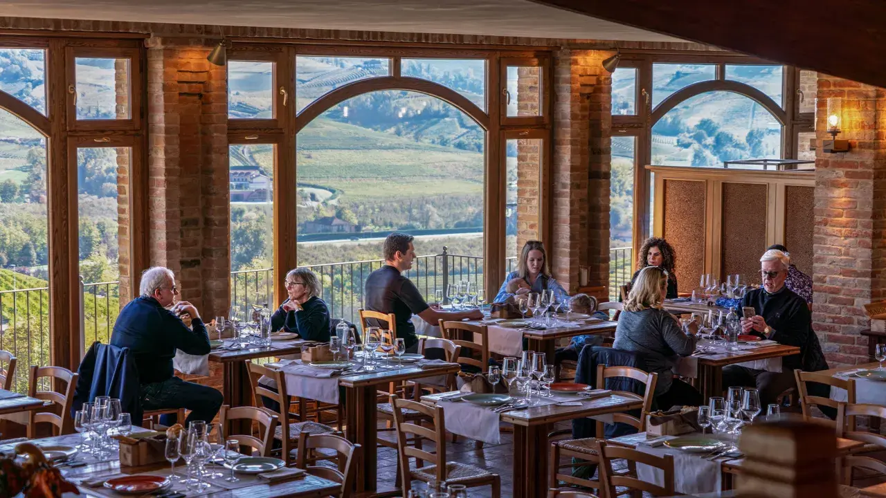 Persone cenano in un ristorante con vista panoramica su un'azienda agricola marrone. Tavoli apparecchiati con cura, mattoni a vista e grandi finestre creano un'atmosfera accogliente.