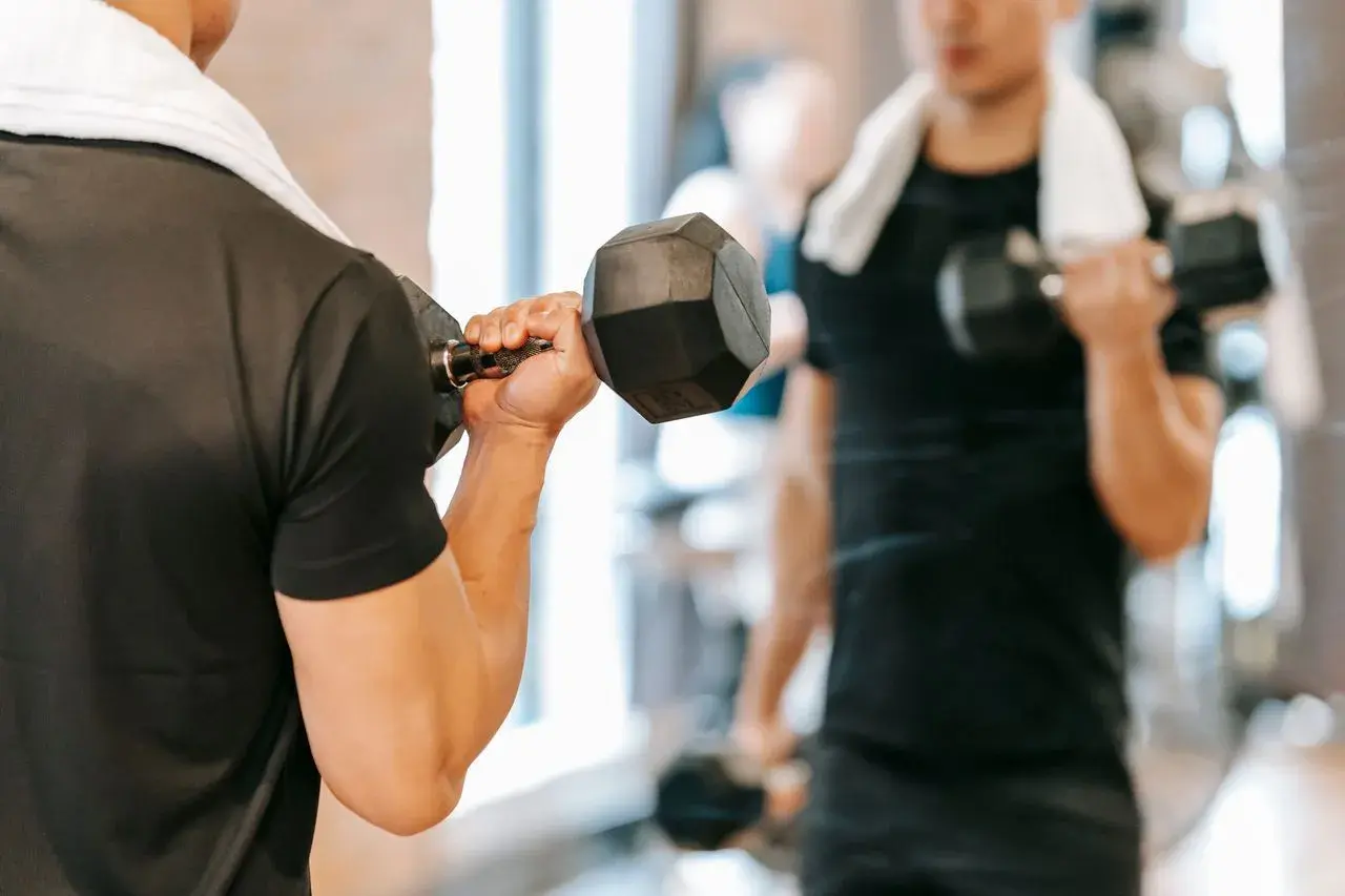 Mężczyzna ćwiczy biceps z hantlem, zaczynając dzień od treningu na czczo. W tle widać jego odbicie w lustrze.