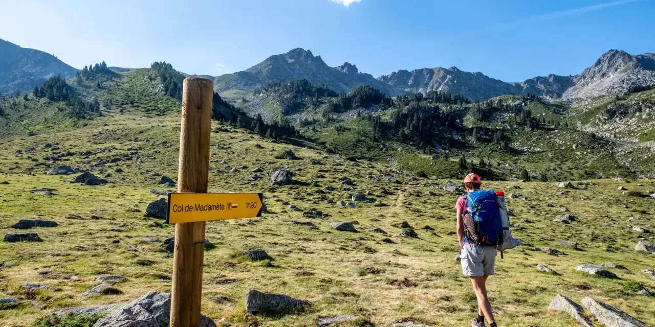 Randonneuse sur le GR10, suivant le panneau jaune indiquant le Col de Madamète. Paysage de montagne verdoyant.