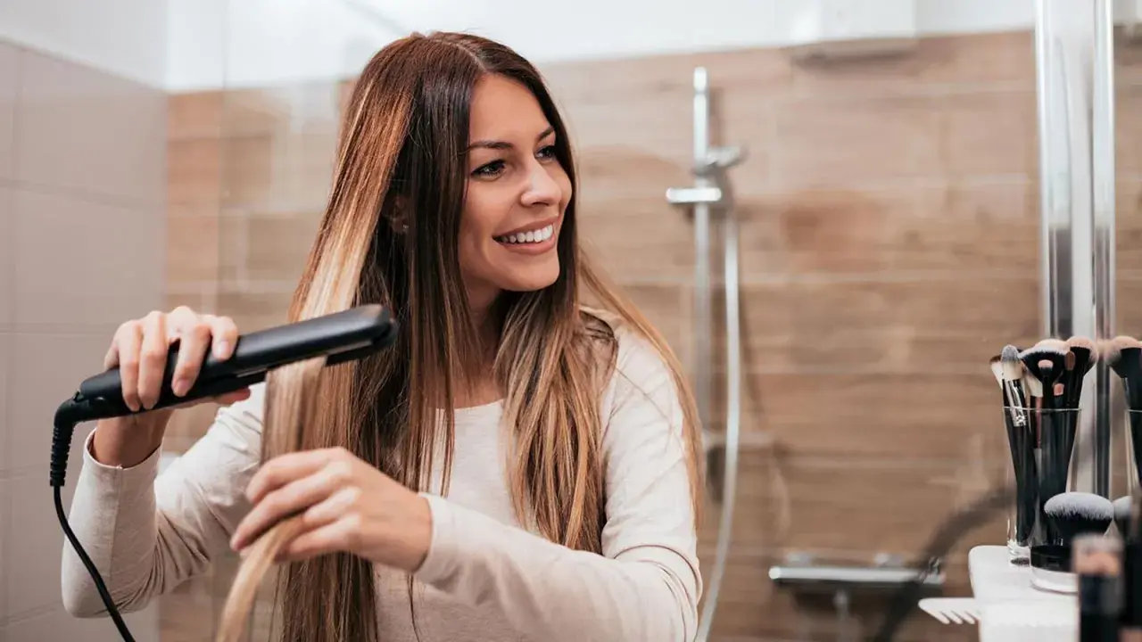 Mujer sonriendo mientras se alisa el pelo con una plancha, buscando el secreto de como alisar el pelo para siempre.