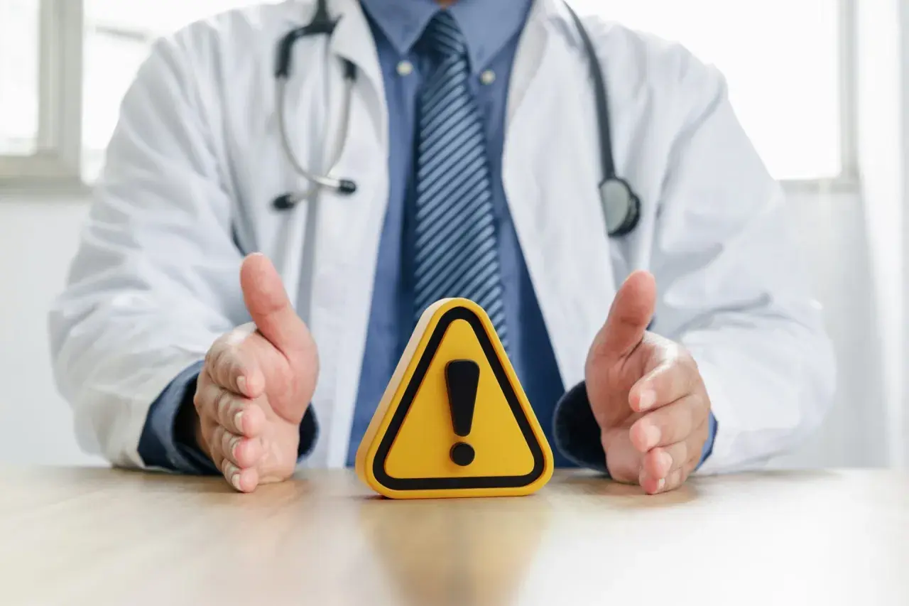Medico con camice bianco e stetoscopio, mani aperte su un segnale di pericolo. Attenzione alle raccomandazioni ministeriali per la salute.