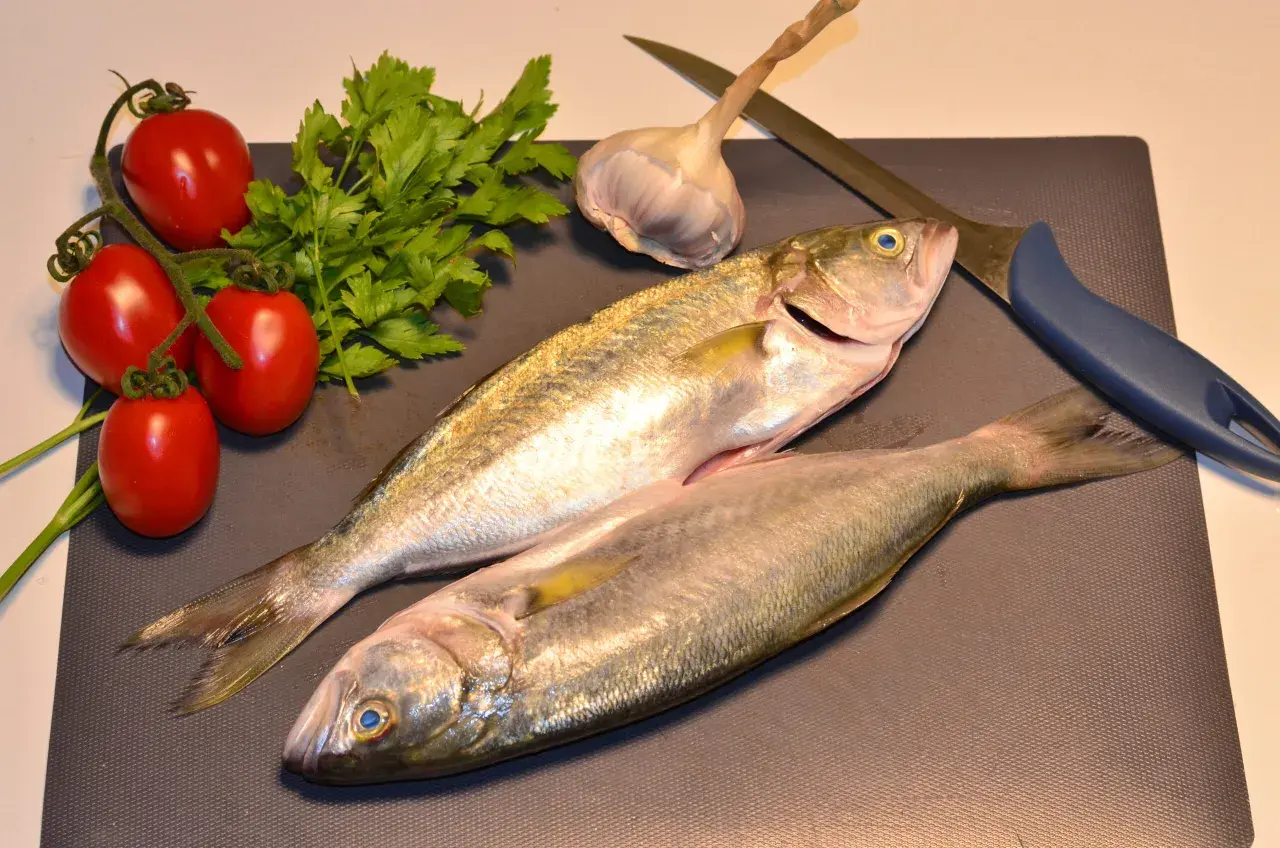 Due pesci serra freschi, pomodorini, prezzemolo, aglio e un coltello su un tagliere, pronti per una cena deliziosa.