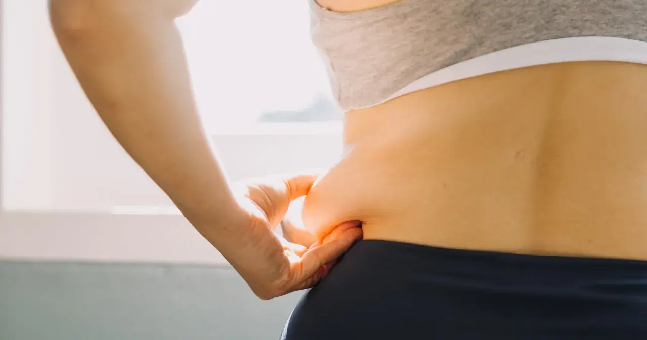 Dłoń chwyta fałd skóry na boku, symbolizując troskę o sylwetkę i chęć poznania, jak przyspieszyć metabolizm.