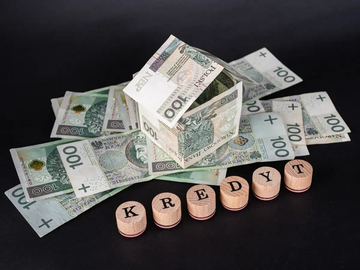 Dom z banknotów 100 zł, symbolizujący kredyt hipoteczny. Słowo "KREDYT" ułożone z drewnianych pieczątek.