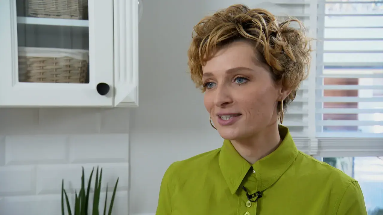 Katarzyna Jaroszyńska, wiek nieznany, w zielonej koszuli, z kręconymi blond włosami, uśmiecha się. W tle biała kuchnia.