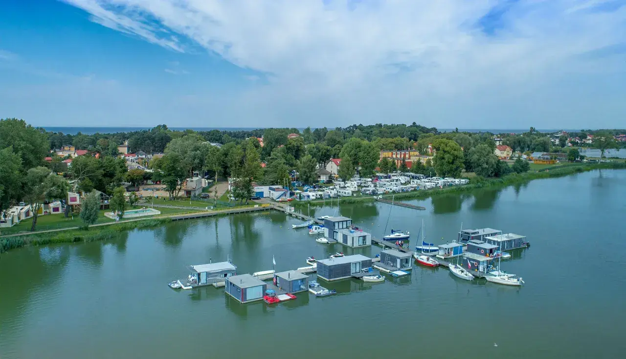 Nowoczesne houseboat'y unoszą się na spokojnej wodzie, obok kempingu z kamperami.