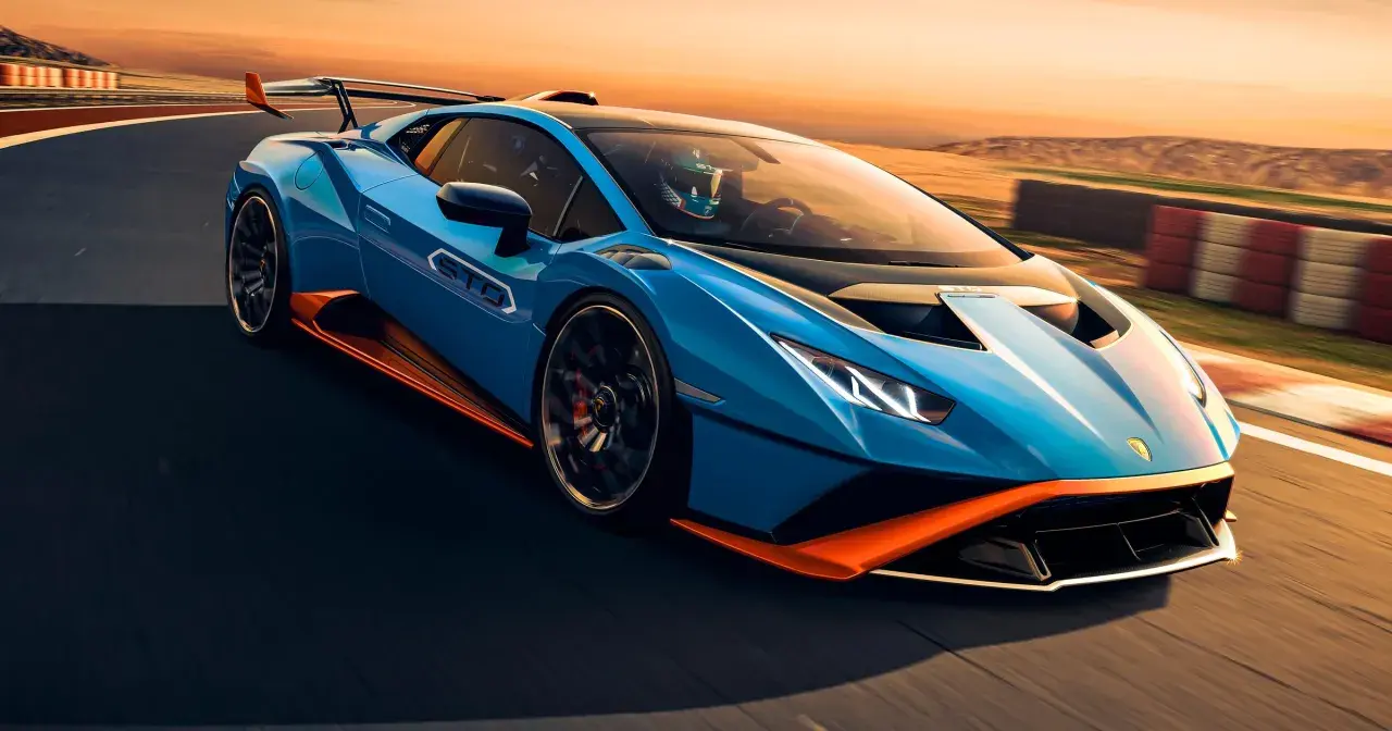 Niebieski Lamborghini Huracan STO z pomarańczowymi akcentami pędzi po torze wyścigowym podczas zachodu słońca.