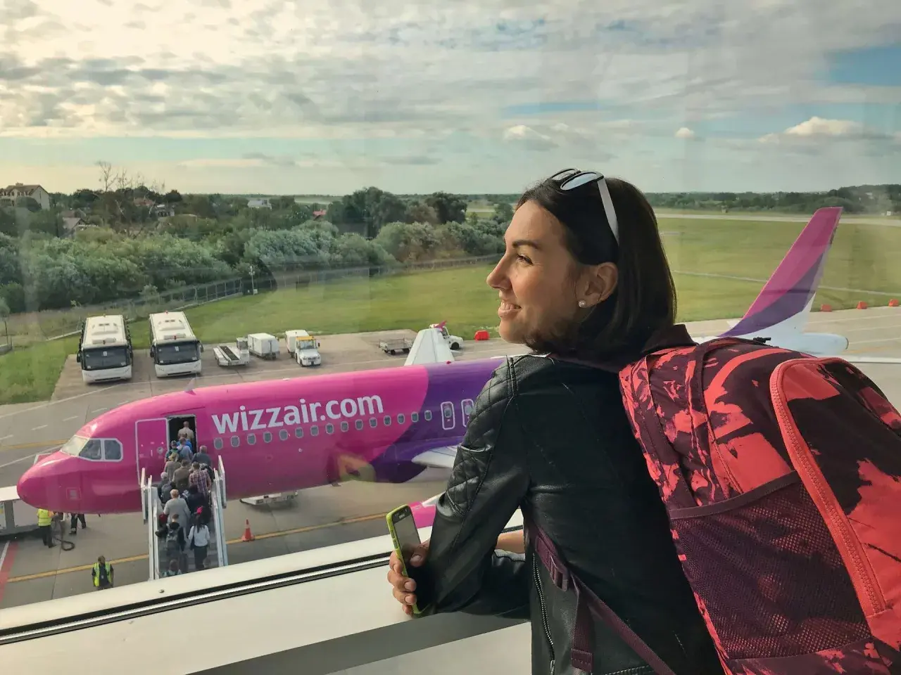 Kobieta z plecakiem i telefonem czeka na lot Wizzair. W tle różowy samolot i pasażerowie wsiadający na pokład.