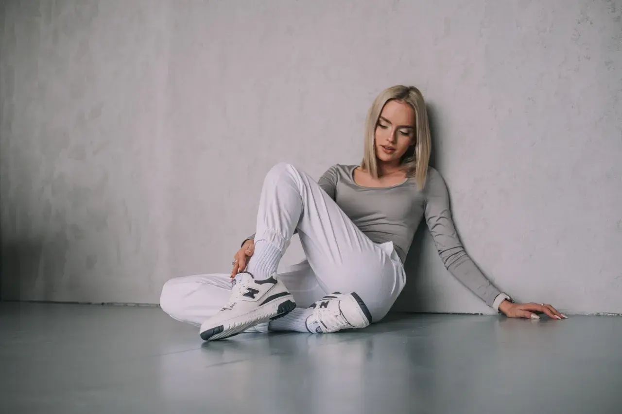 Blondynka w białych spodniach i szarym topie siedzi na podłodze, opierając się o ścianę. Na nogach ma stylowe snekarsy.