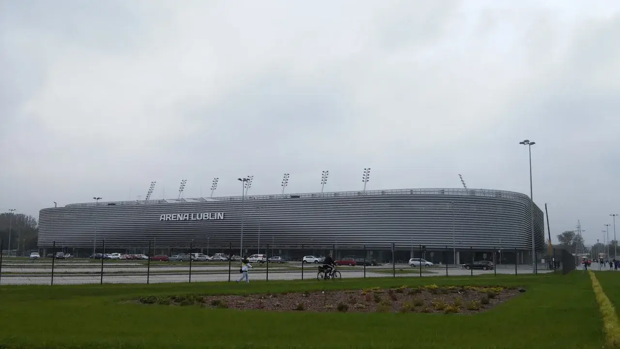 Nowoczesna Arena Lublin z napisem "ARENA LUBLIN" na fasadzie. Obok parking, a na pierwszym planie trawnik. Pojemność obiektu robi wrażenie.