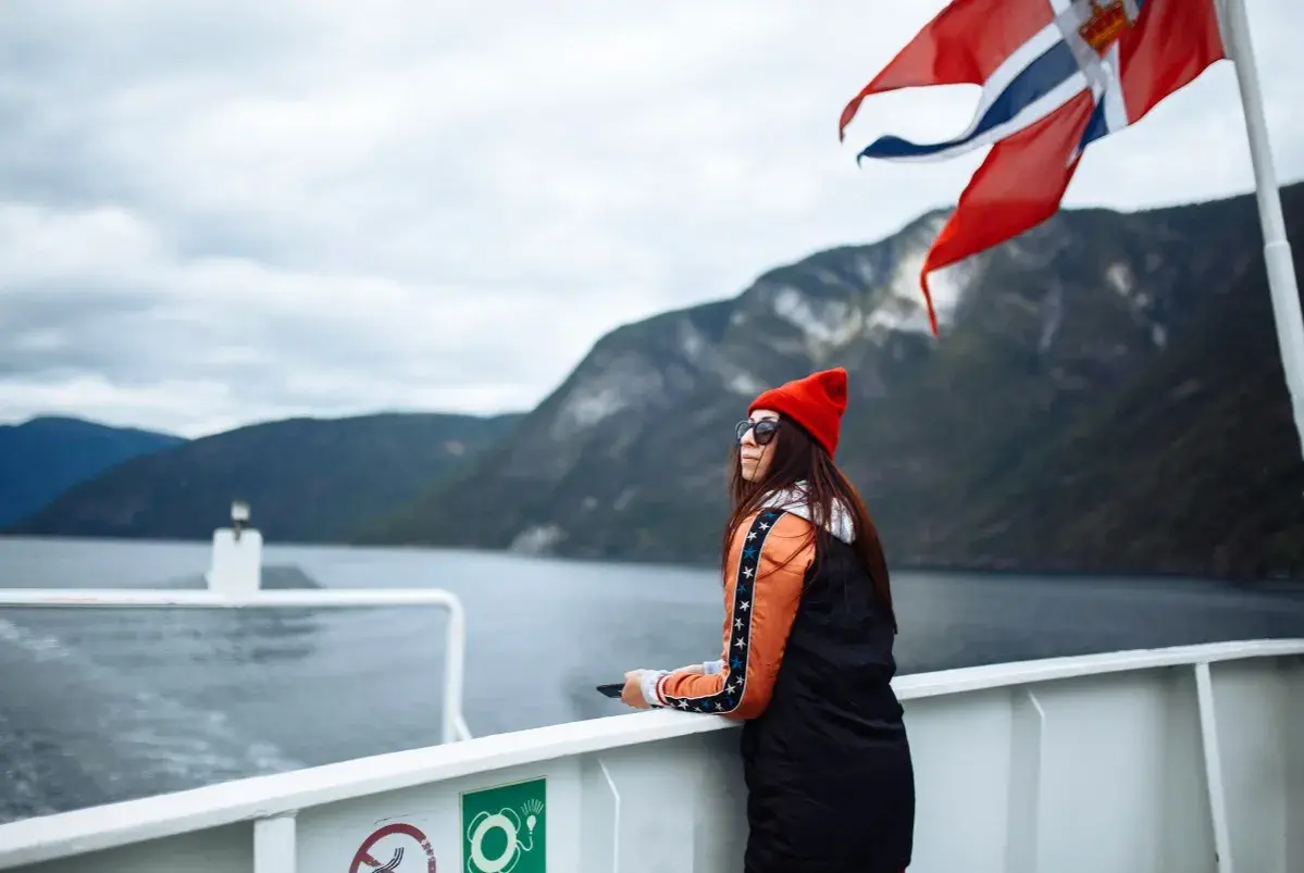 Kobieta na statku pod norweską flagą podziwia fiordy. Podróż po Norwegii, mimo że piękna, może wiązać się z wysokimi cenami.