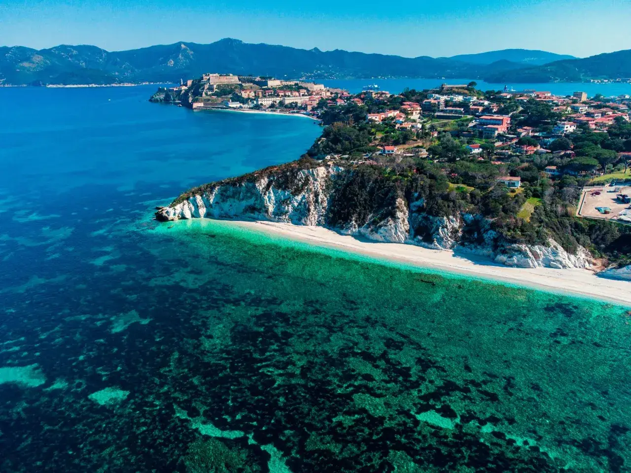 Arcipelago Toscano: una costa con scogliere bianche, spiaggia dorata e mare cristallino, con un borgo storico in lontananza. Un gioiello tra le isole visitabili.