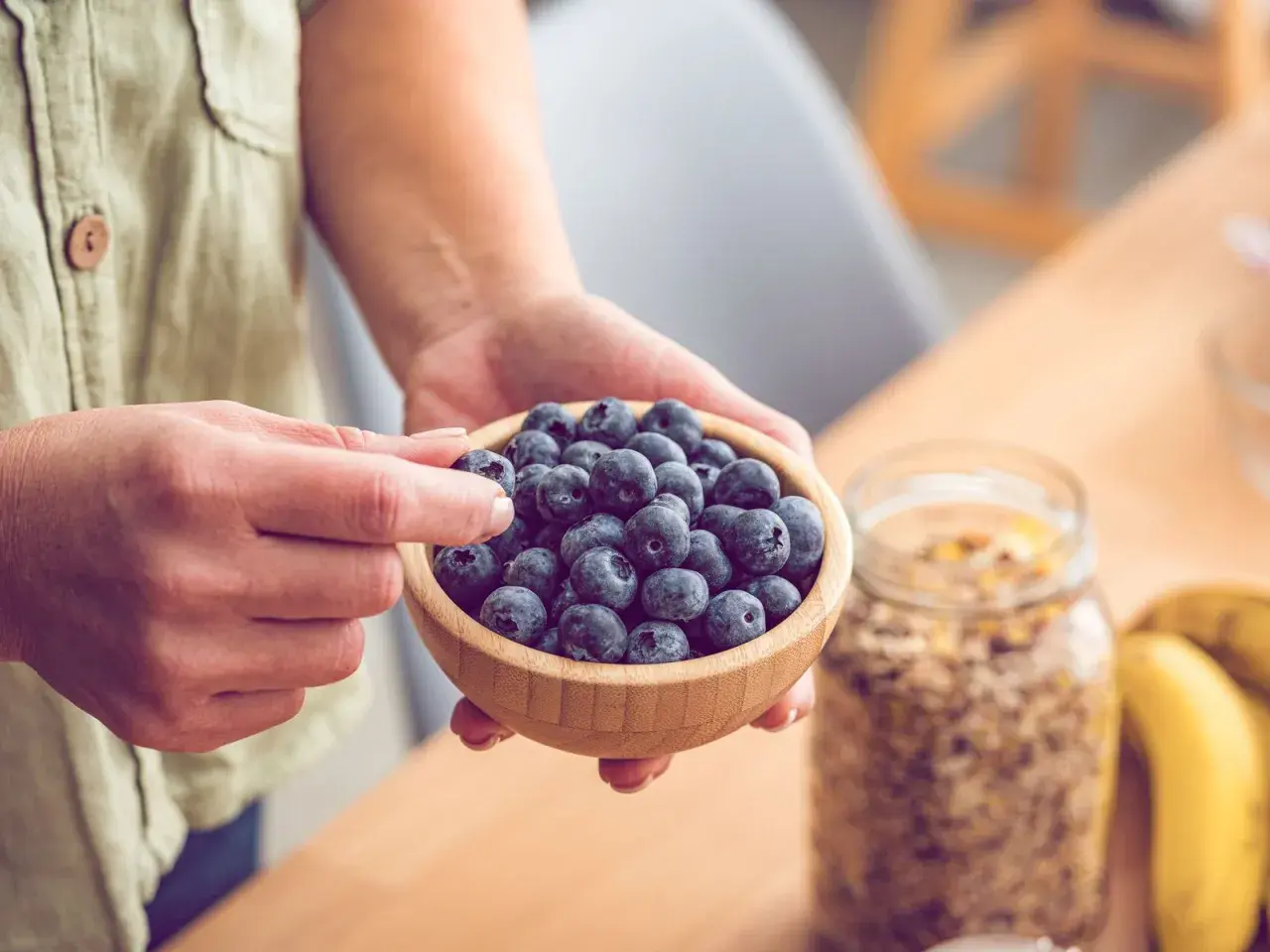 Hände halten eine Schale voller Blaubeeren, reich an Flavonoiden. Daneben stehen Müsli und Bananen – gesunde Flavonoide Lebensmittel für den Start in den Tag.