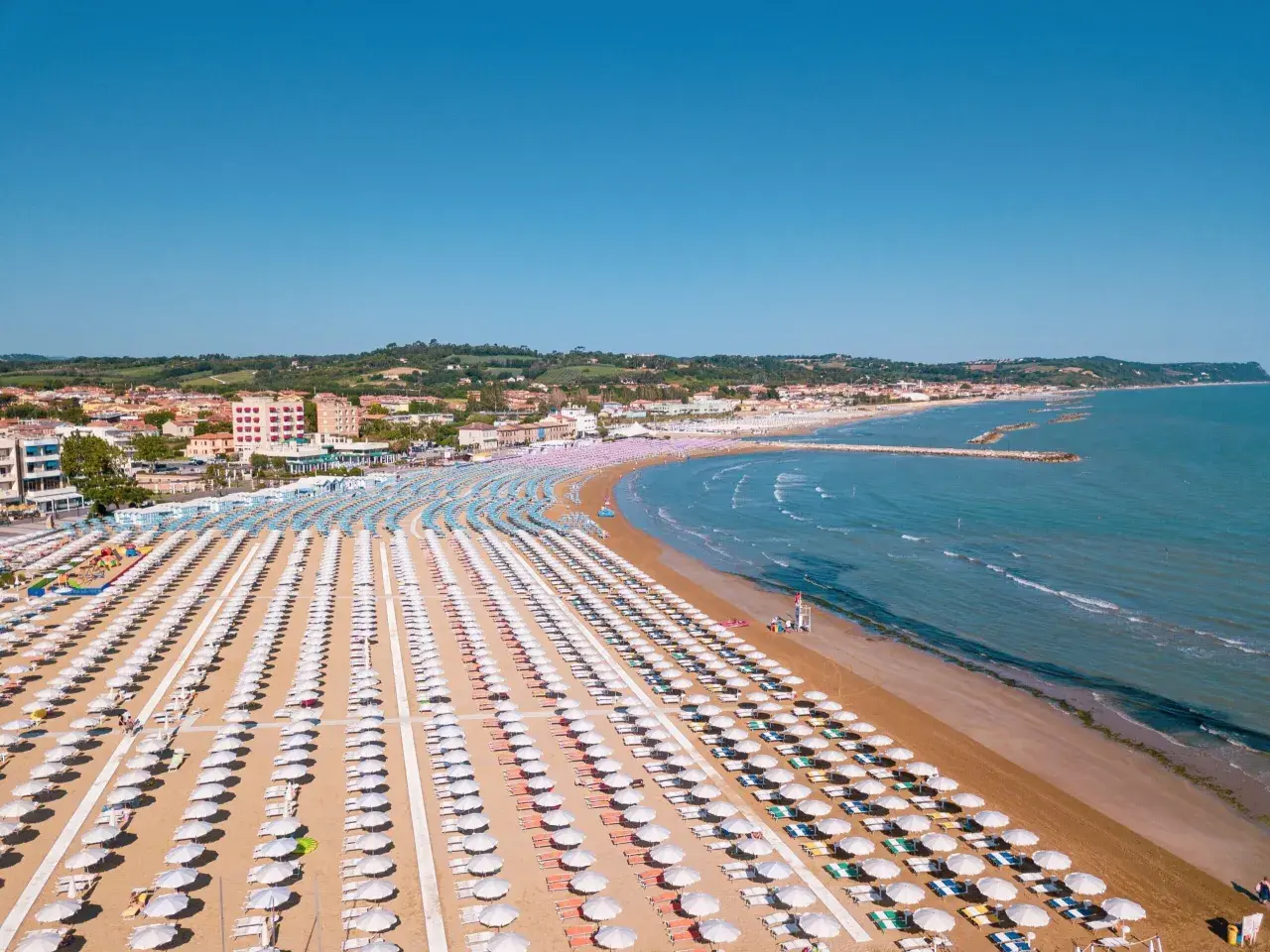 Fano mare: file ordinate di ombrelloni bianchi e colorati sulla sabbia dorata, con il mare azzurro e la costa sullo sfondo.