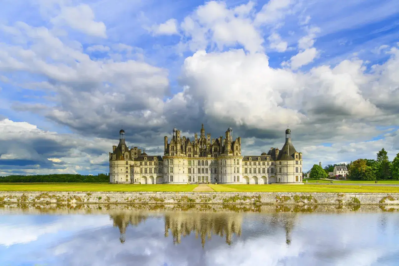 Zamek Chambord nad Loarą odbijający się w wodzie, pod burzliwym, błękitnym niebem.