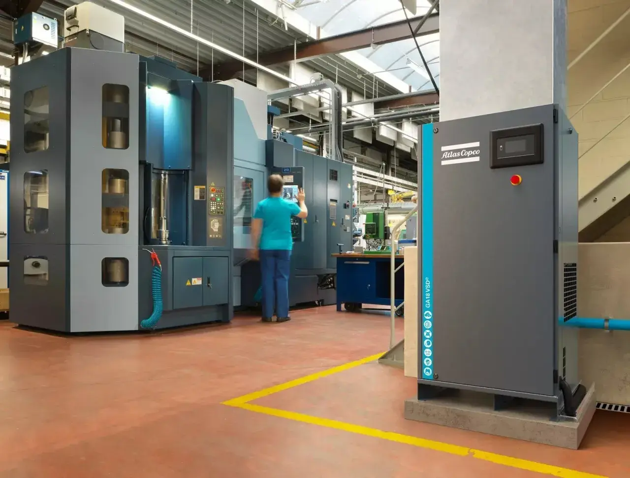 En una fábrica, un operario interactúa con una máquina industrial. A la derecha, un compresor Atlas Copco, que puede ser con aceite o sin aceite, espera su turno.