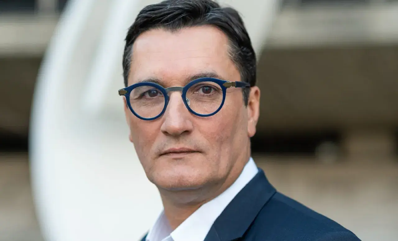 Olivier Dartigolles, un homme aux lunettes bleues, pose pour un portrait. Il est vêtu d'un costume sombre, évoquant une image de famille et de professionnalisme.