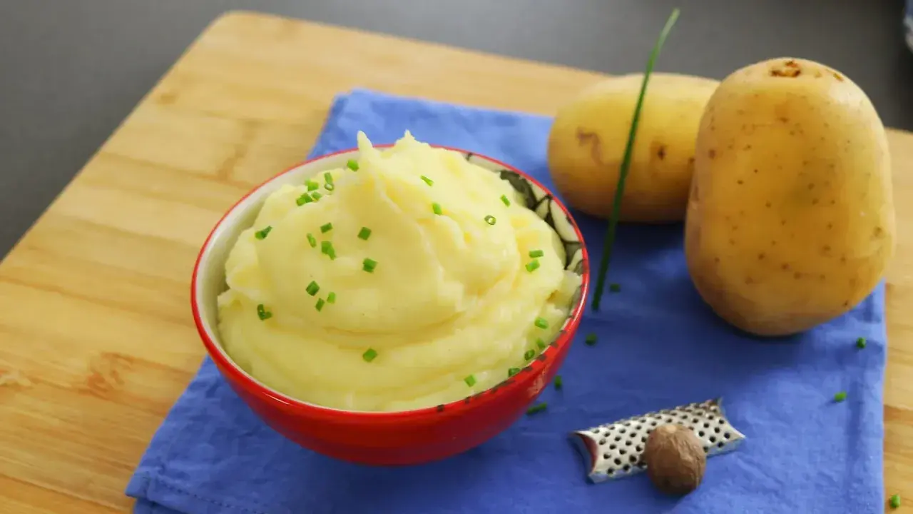 Un cremoso puré de patatas, adornado con cebollino fresco, junto a patatas crudas y una nuez moscada.