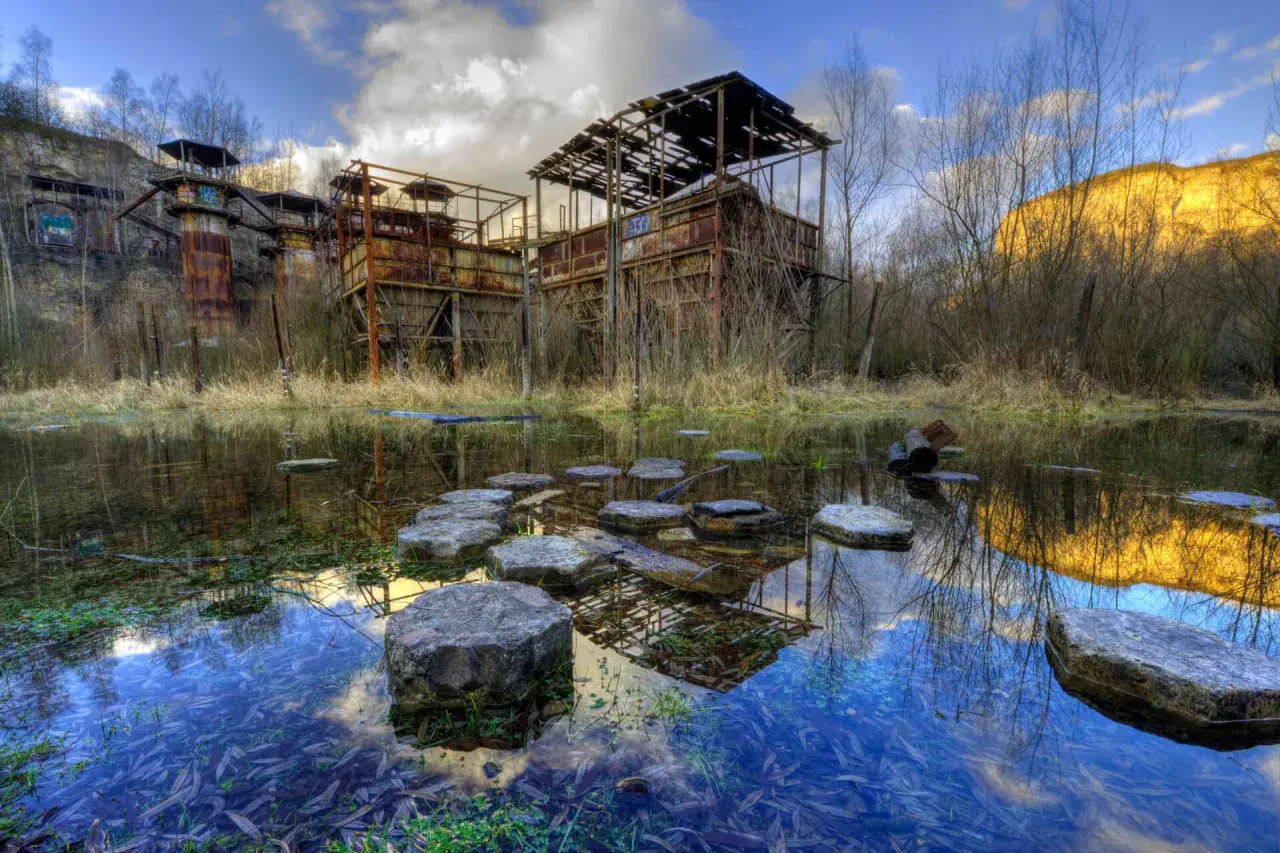 Zrujnowana fabryka, przypominająca opuszczoną "Fabbrica di Schindler", zardzewiałe konstrukcje i stawy odbijające niebo.