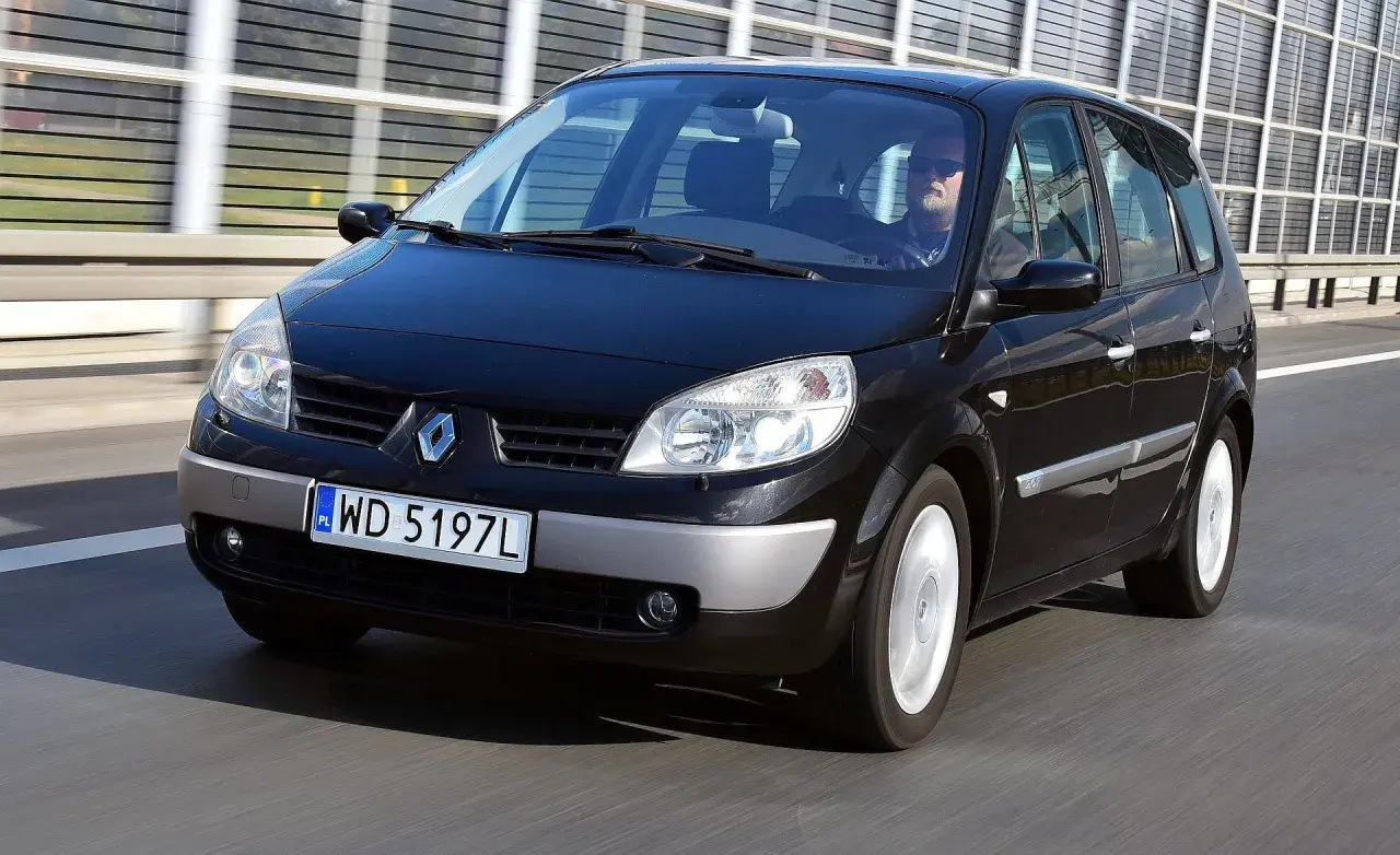 Czarny Renault Scenic 2 jedzie po drodze. Kierowca w okularach przeciwsłonecznych prowadzi auto.