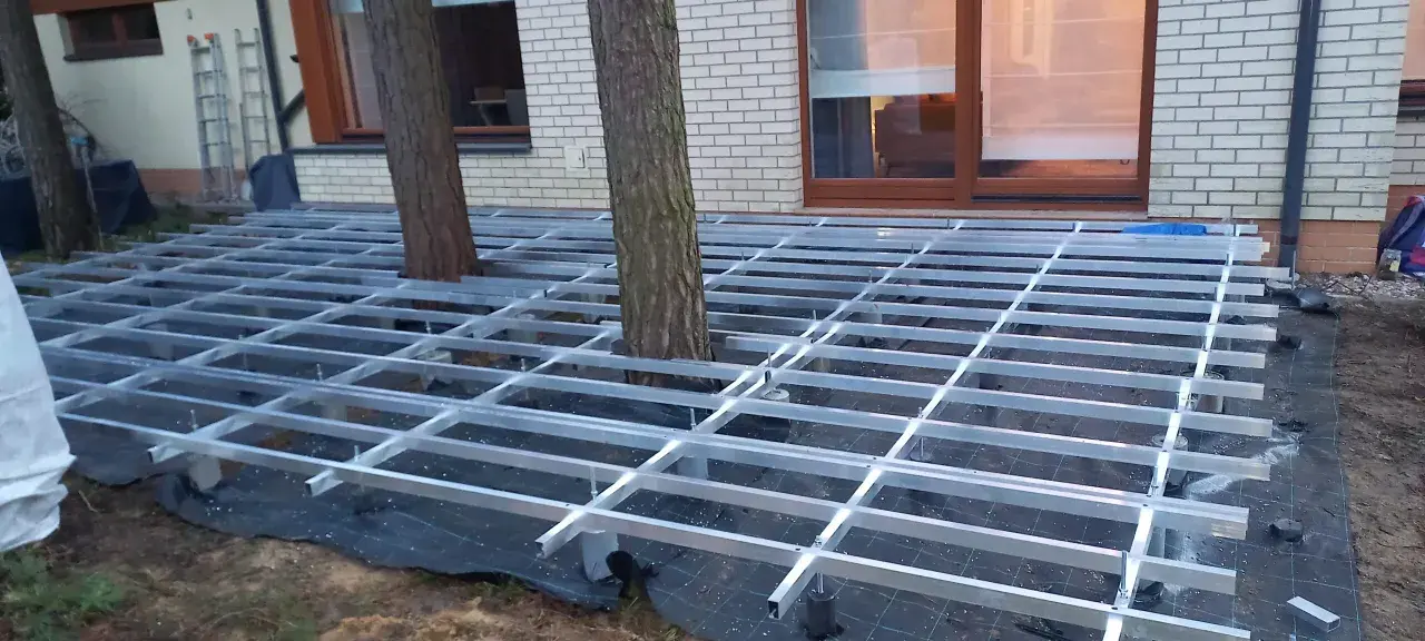 Trwa układanie deski kompozytowej na aluminiowym stelażu, który omija drzewa rosnące na tarasie.