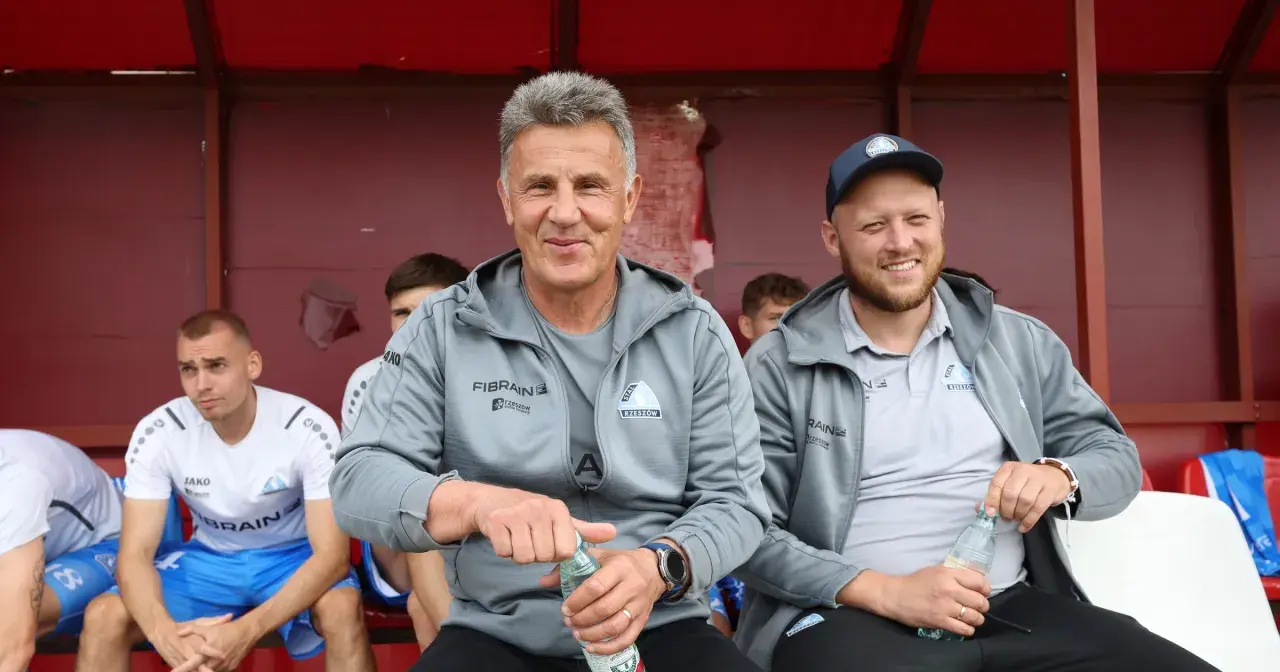 Marek Zub, trener Stali Rzeszów, z uśmiechem na twarzy, siedzi na ławce z butelką wody.