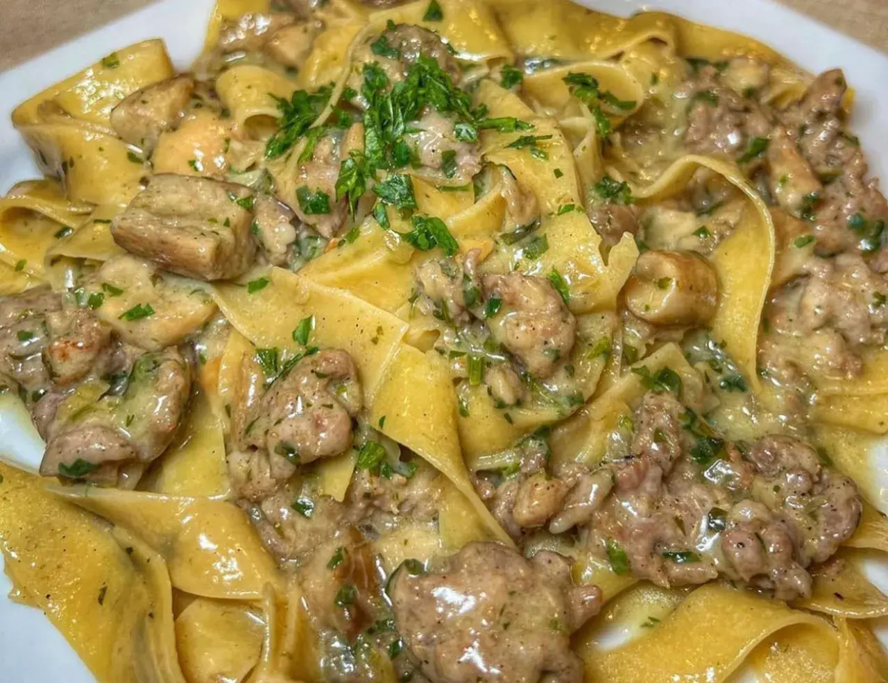 Pappardelle cremose con salsiccia e funghi, guarnite con prezzemolo fresco. Un piatto ricco e saporito.