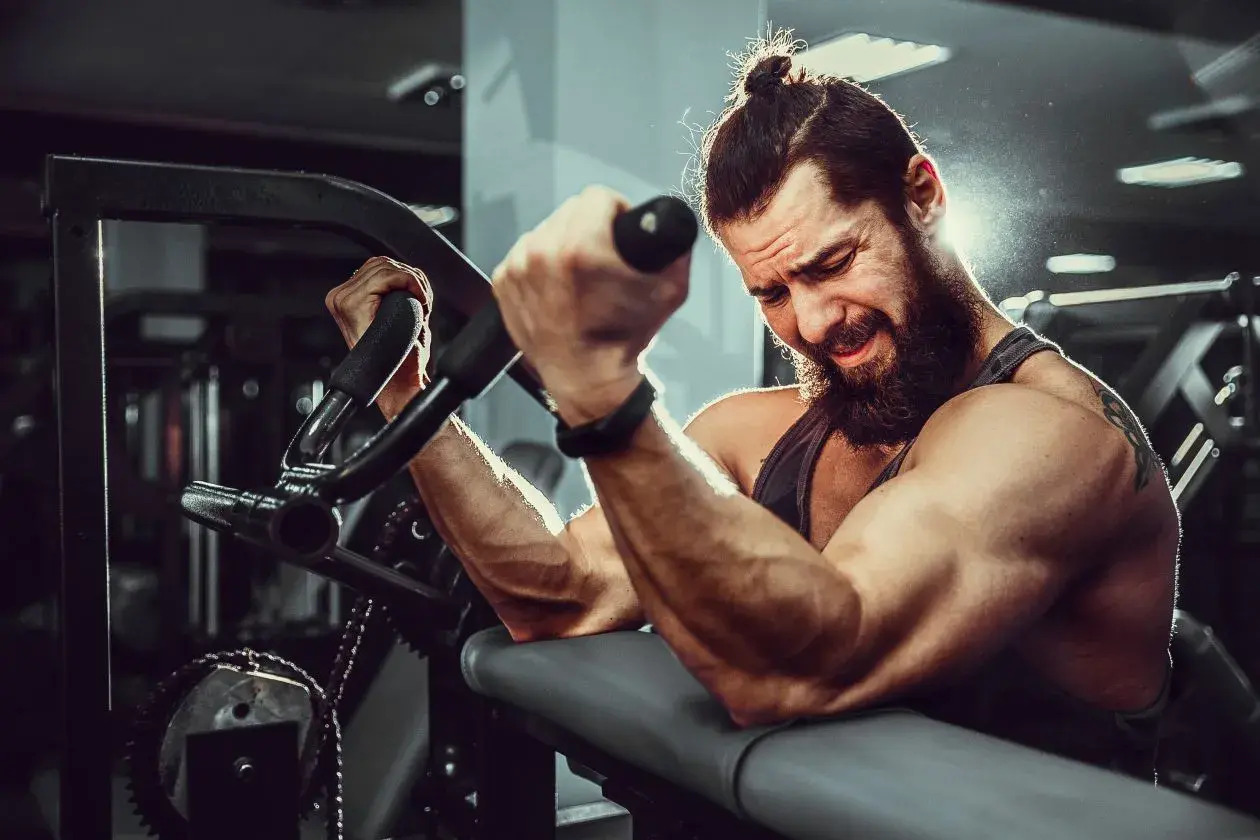 Mężczyzna z brodą i kitką ćwiczy biceps na maszynie. Widać jego napięte mięśnie i determinację. To idealne miejsce, by budować **mutant mass sklad**.