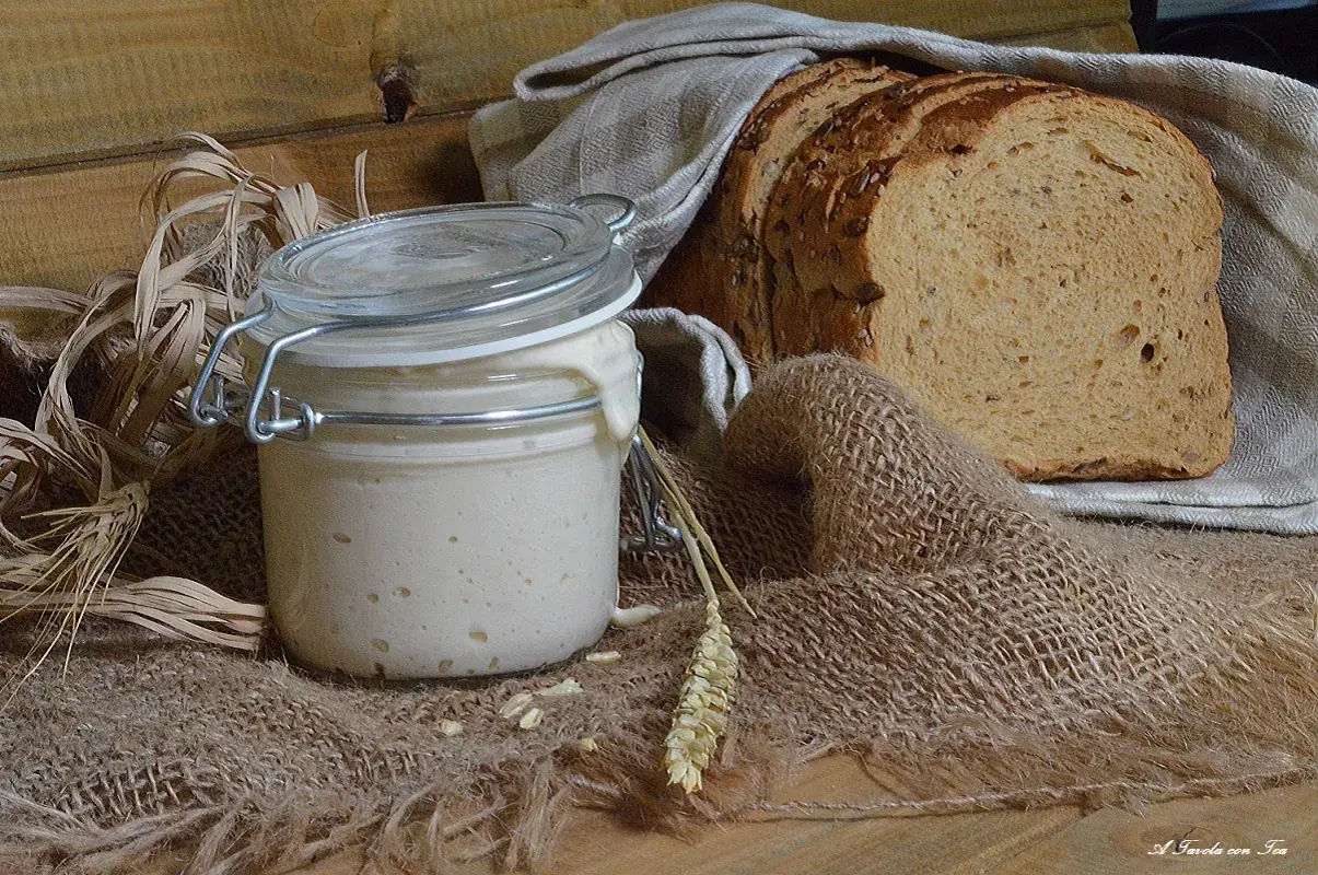 Un barattolo di lievito madre attivo, con una fetta di pane rustico sullo sfondo. Ingredienti naturali per un pane fragrante.
