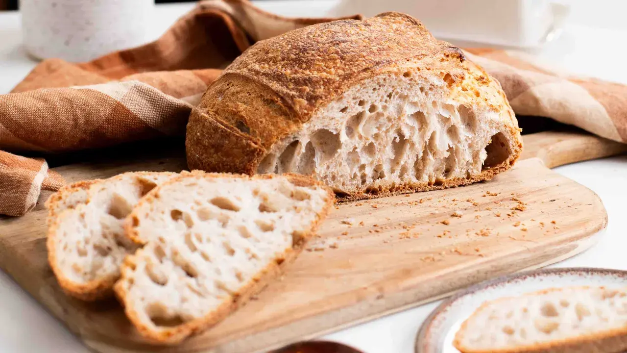 Un delicioso pan de masa madre, con su corteza dorada y miga aireada, reposa sobre una tabla de madera junto a unas rebanadas.