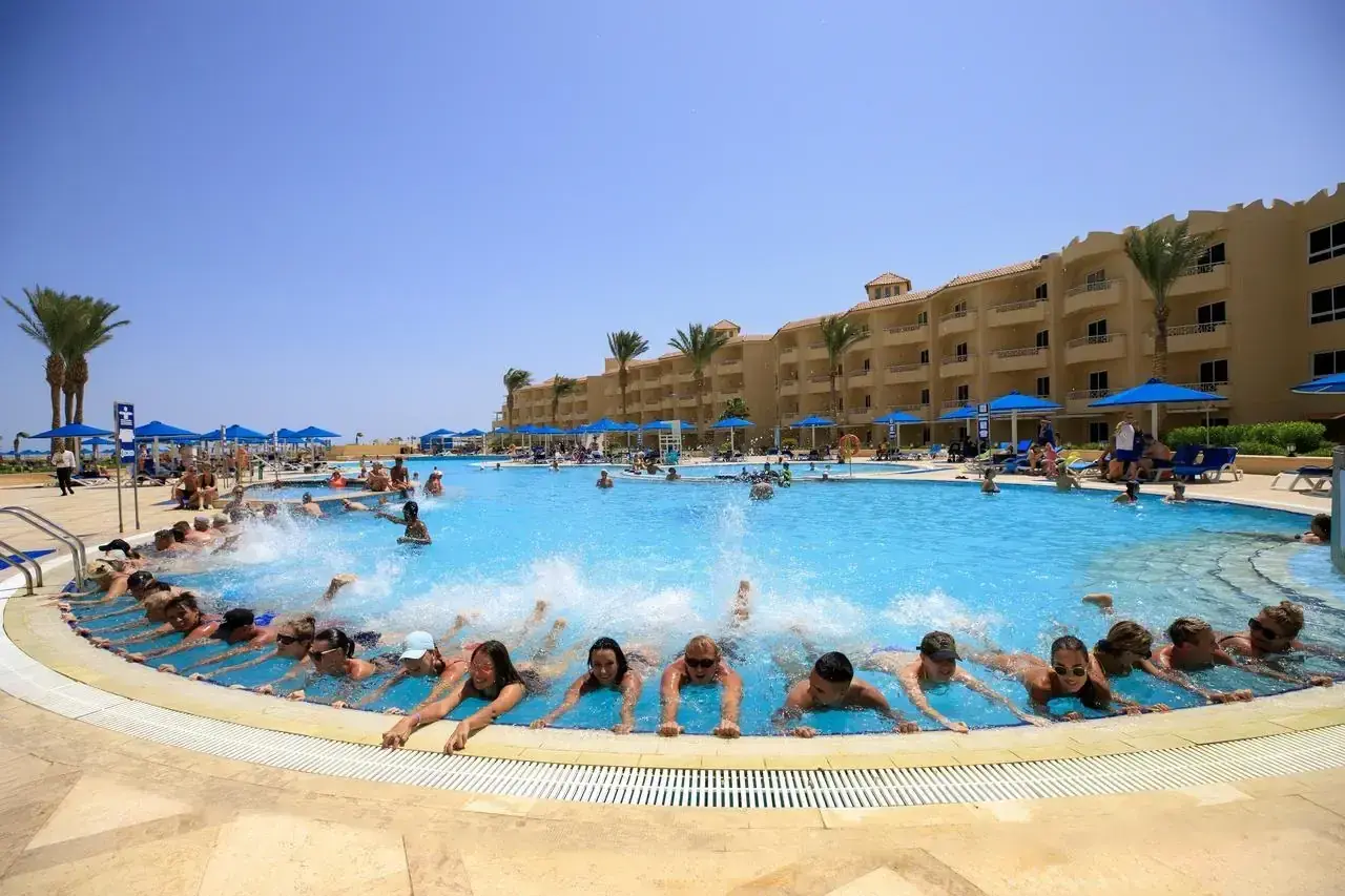 Grupa ludzi pływa w basenie hotelu, tworząc falę. W tle budynek hotelu i palmy. Wiele pozytywnych amwaj blue beach resort & spa opinie potwierdza ten widok.